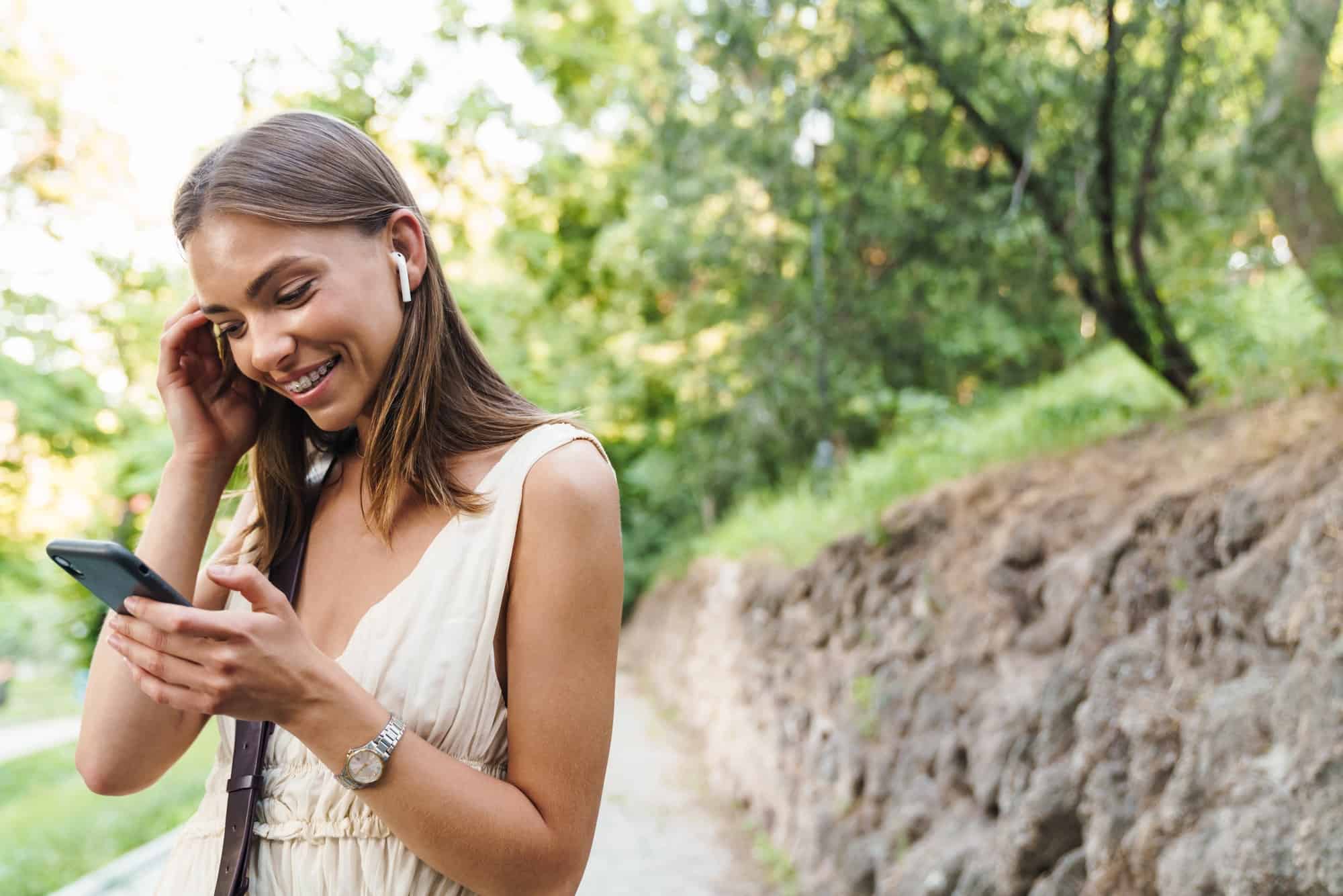 Image of young woman in earbuds laughing and holding cellphone in park