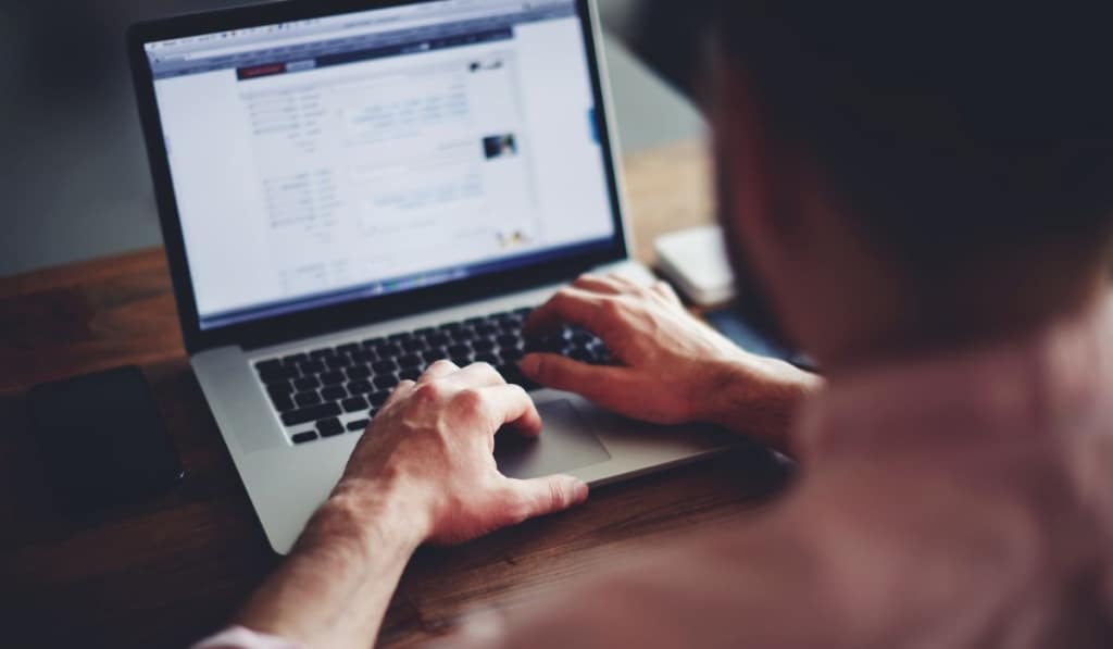 Young male student texting on computer sitting at wooden table 