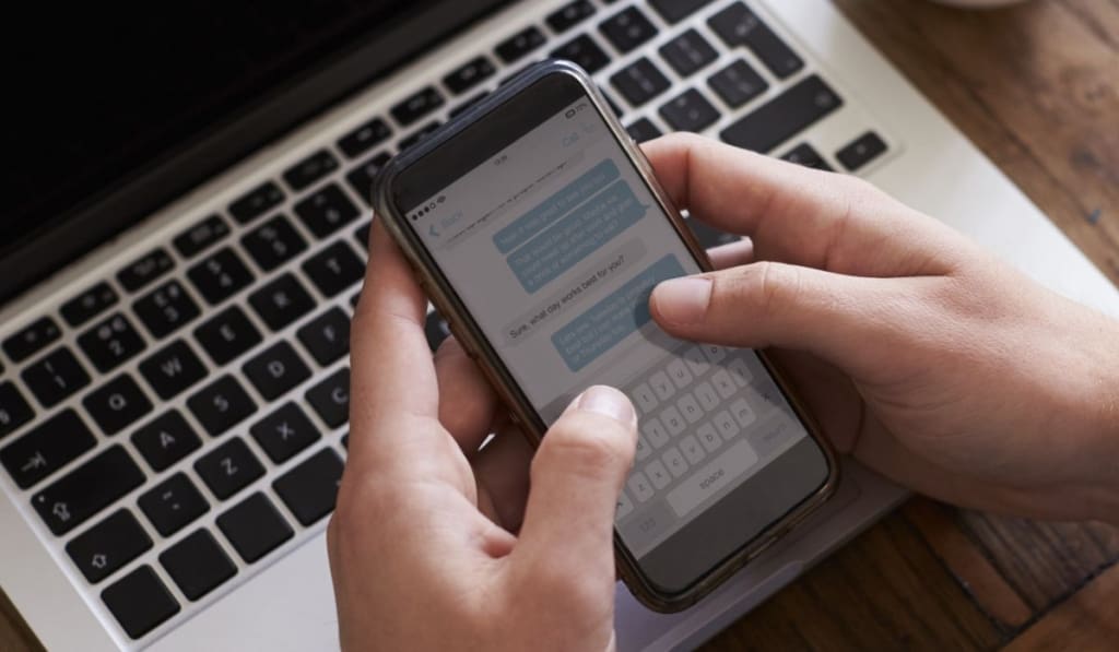 Man Sending Text Message On Mobile Phone Whilst Using Laptop