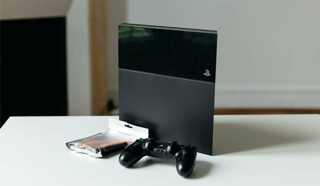 PS4 sitting vertically on a table with a controller and bag of batteries